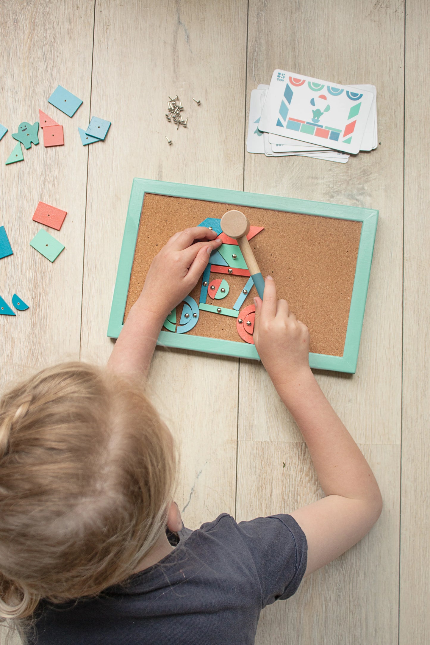Child playing with a wooden puzzle on a cork board with scattered pieces on a wooden floor.